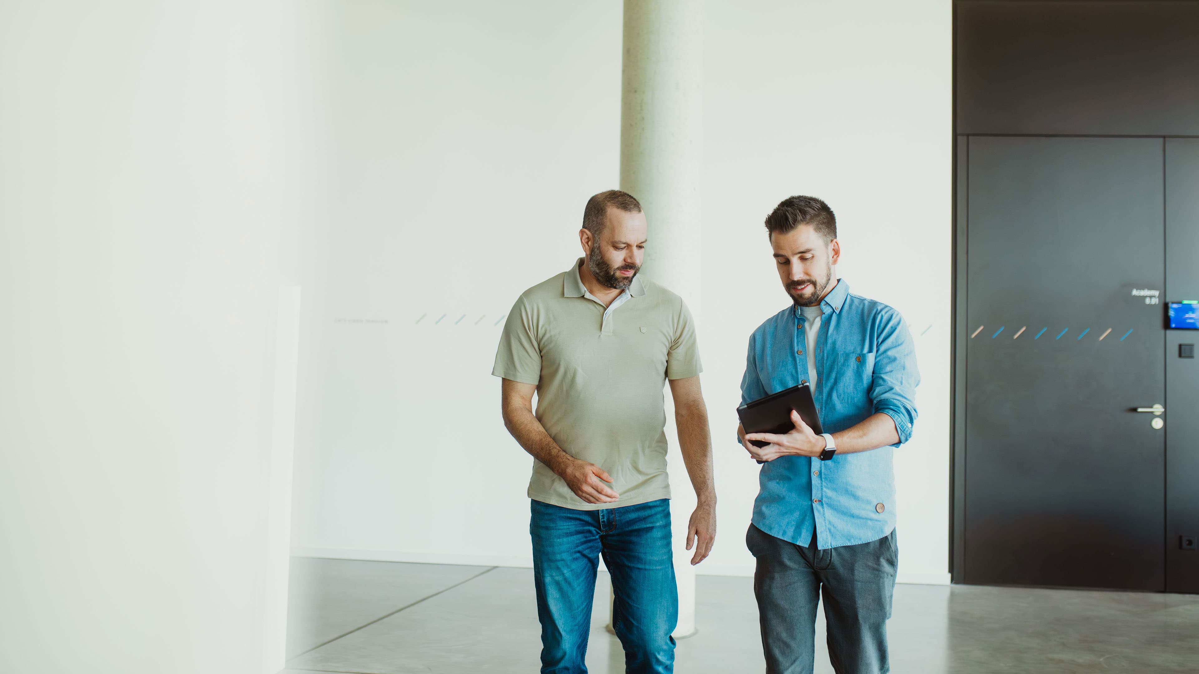 Mobile Device Management: Zwei Mitarbeiter gehen durch ein Büro, während einer ein Tablet nutzt.