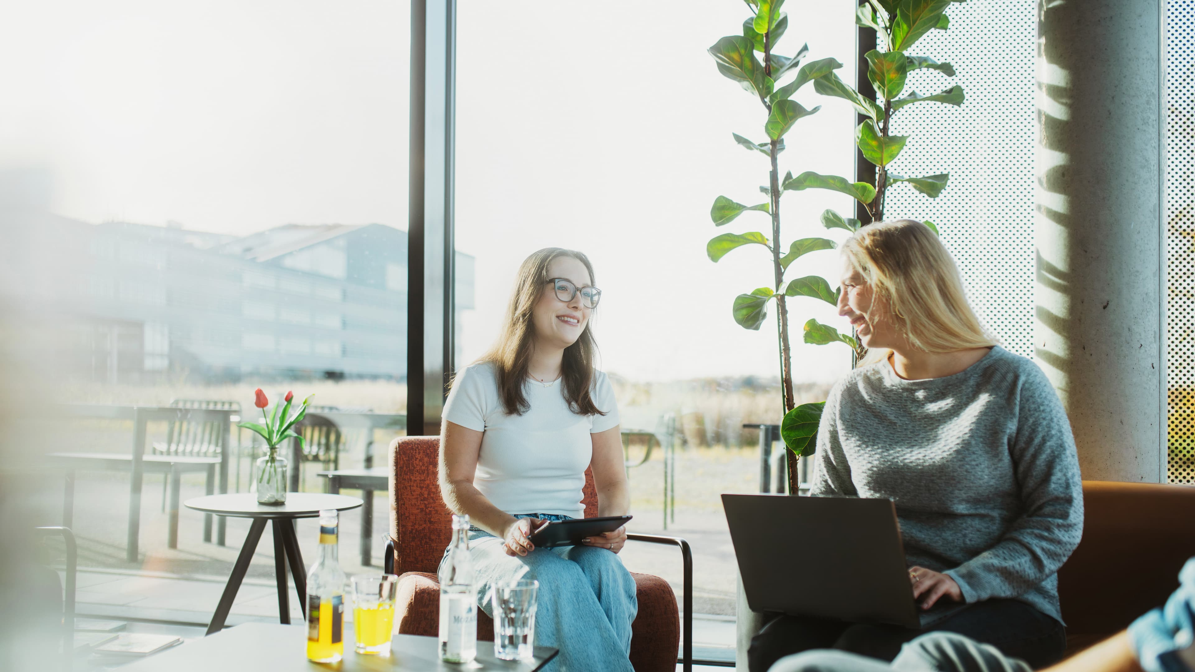 Two women are sitting in a brightly lit office area; one is holding a tablet, and the other is using a laptop.