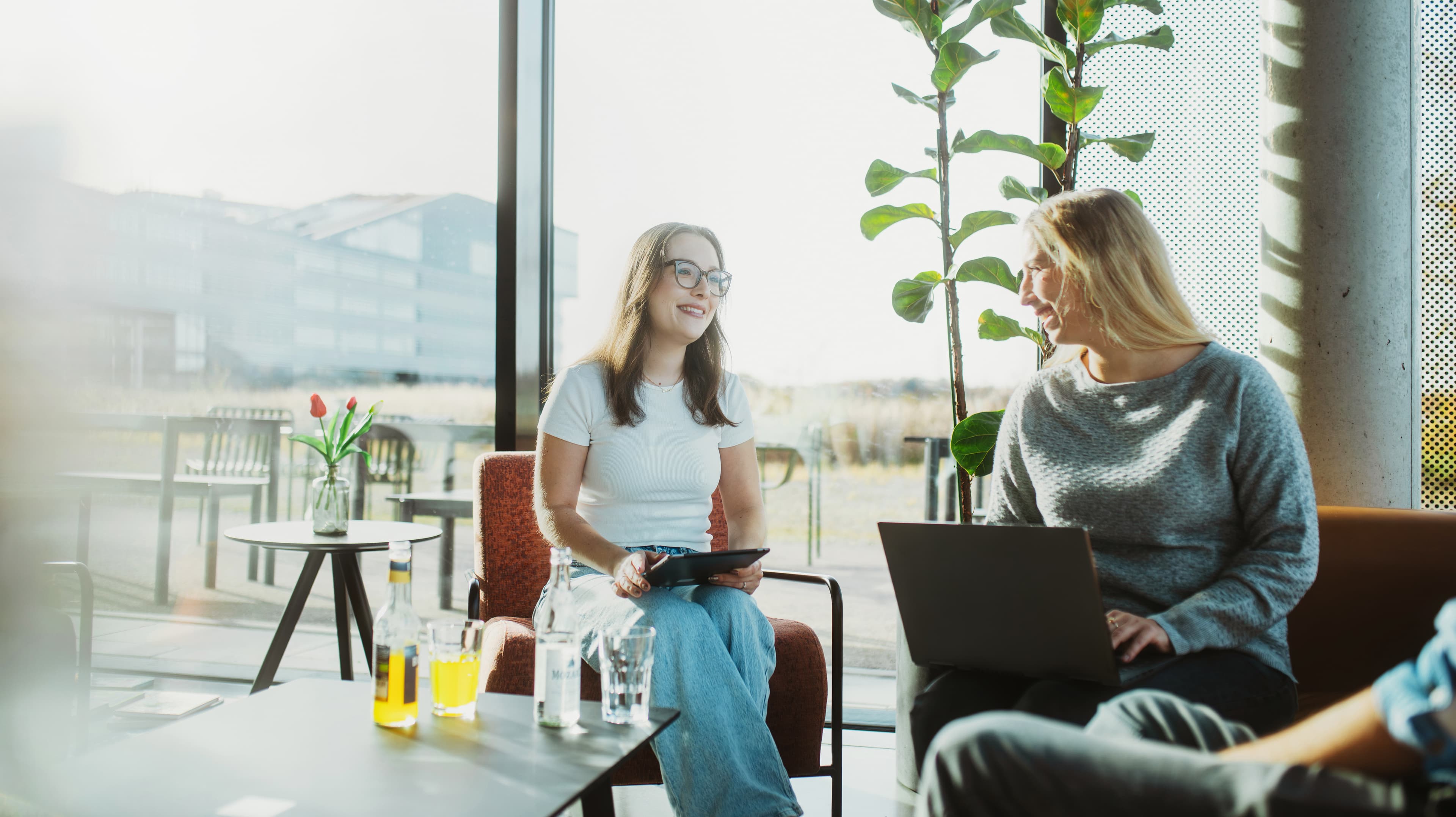 Two women are sitting in a well-lit office, working on a tablet and a laptop.