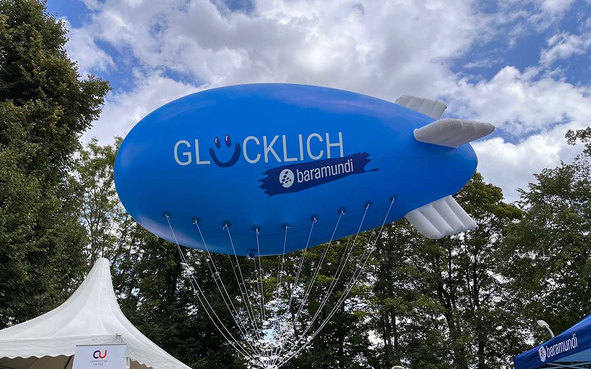 Ein blauer Zeppelin mit der Aufschrift "GLÜCKLICH" und dem Logo von baramundi schwebt über einem Veranstaltungszelt im Freien