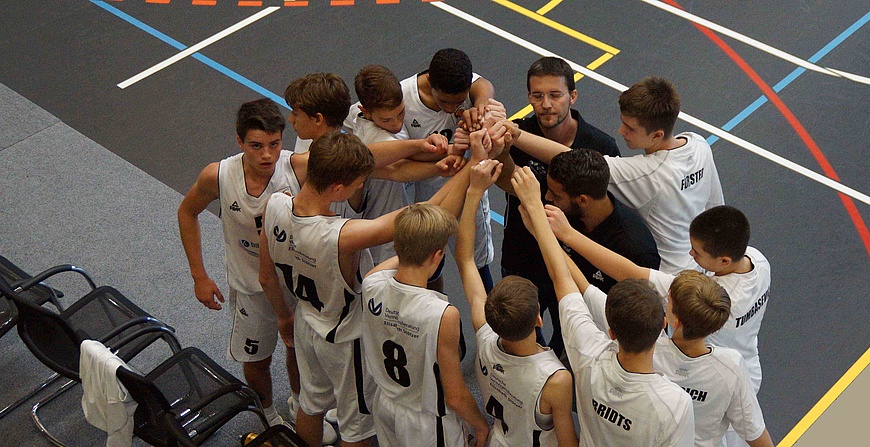 Mehrere Kinder im Trikot eines Basketballteams bilden einen Kreis und legen ihre Hände aufeinander, während ein Trainer in der Mitte steht