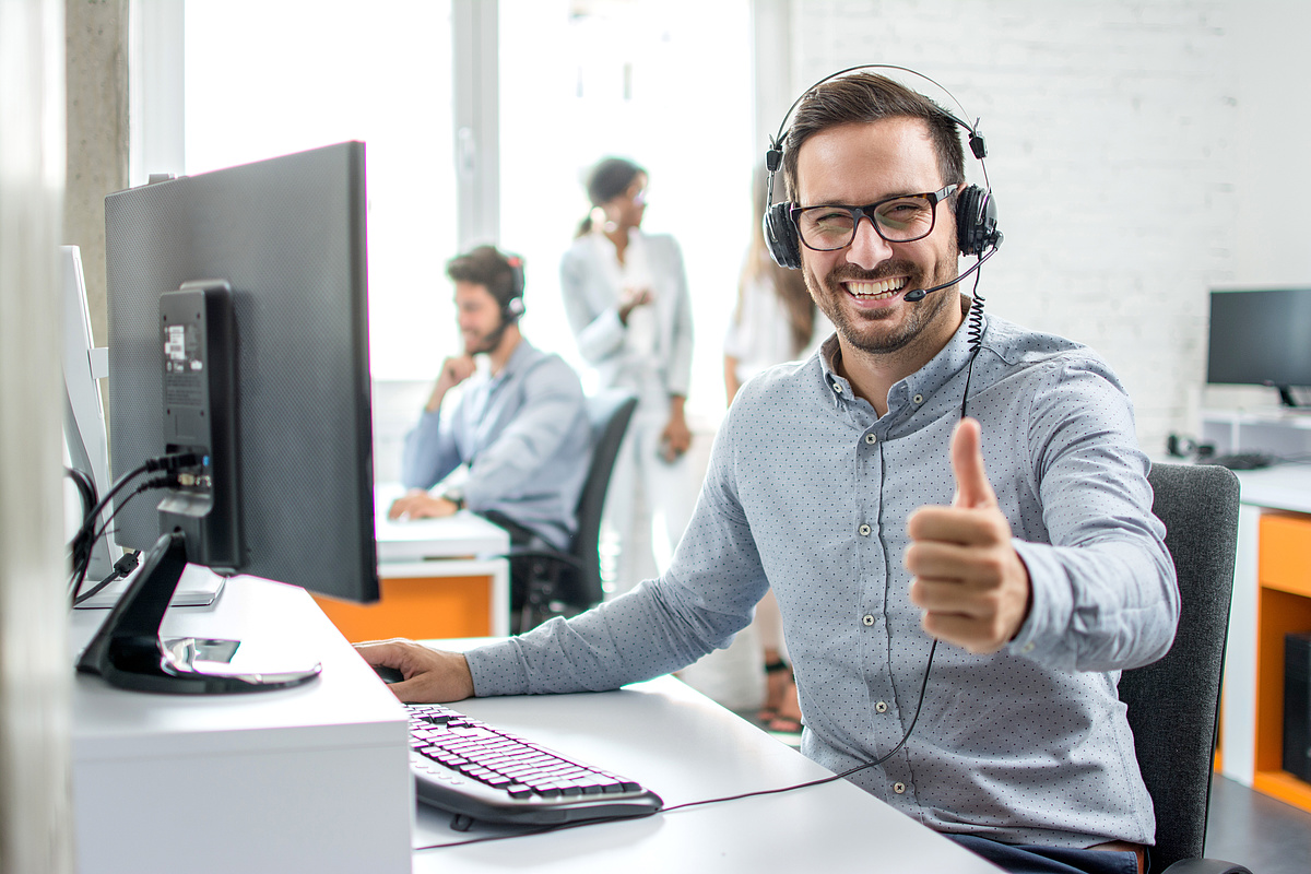 Mitarbeiter im Büro trägt ein Headset und zeigt Daumen hoch vor einem Computer