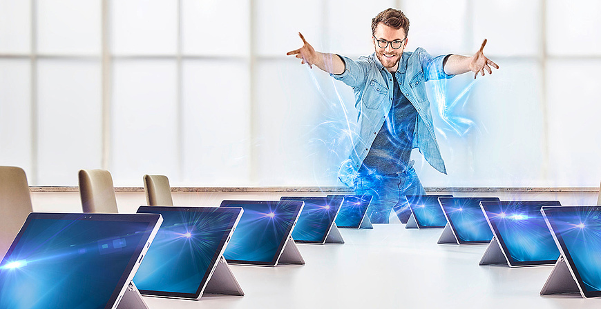 Lächelnder Mann mit ausgebreiteten Armen steht vor einer Reihe von Laptops in einem Konferenzraum