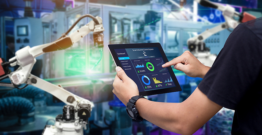 Eine Person bedient ein Tablet mit einer Dashboard-Anzeige in einer industriellen Umgebung mit Roboterarmen im Hintergrund