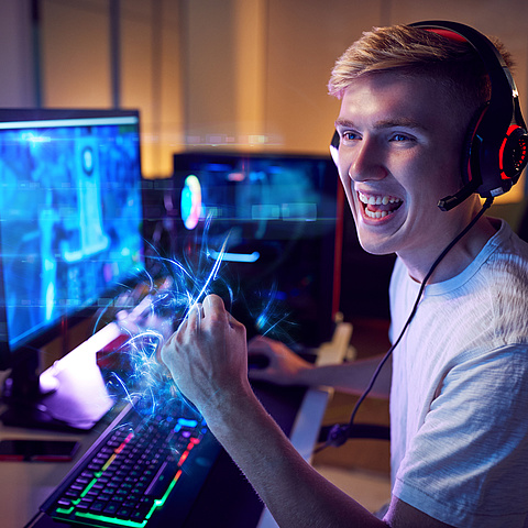 Ein junger Mann sitzt begeistert vor einem Computerbildschirm, während er ein Headset trägt und seine Faust triumphierend in die Luft reckt