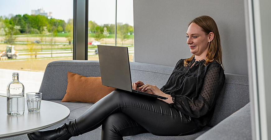 Eine baramundi Mitarbeiterin arbeitet entspannt auf einem Sofa mit einem Laptop im modernen Büro