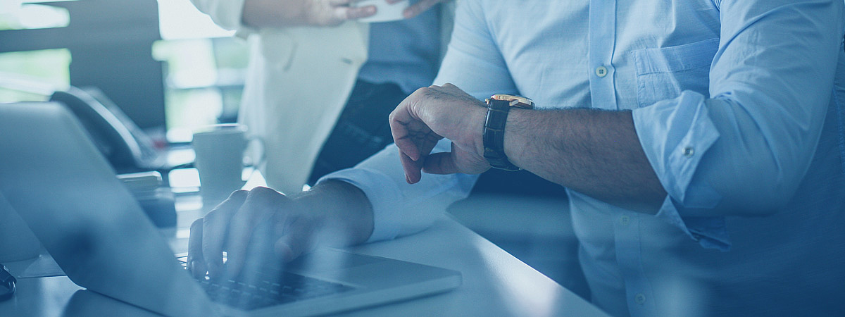 Eine Person arbeitet am Laptop und schaut auf ihre Armbanduhr, während im Hintergrund eine weitere Person steht