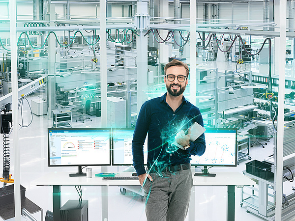 Ein Mann steht in einem modernen industriellen Umfeld mit einem Tablet in der Hand und zwei Bildschirmen vor sich, die die baramundi Management Suite zeigen
