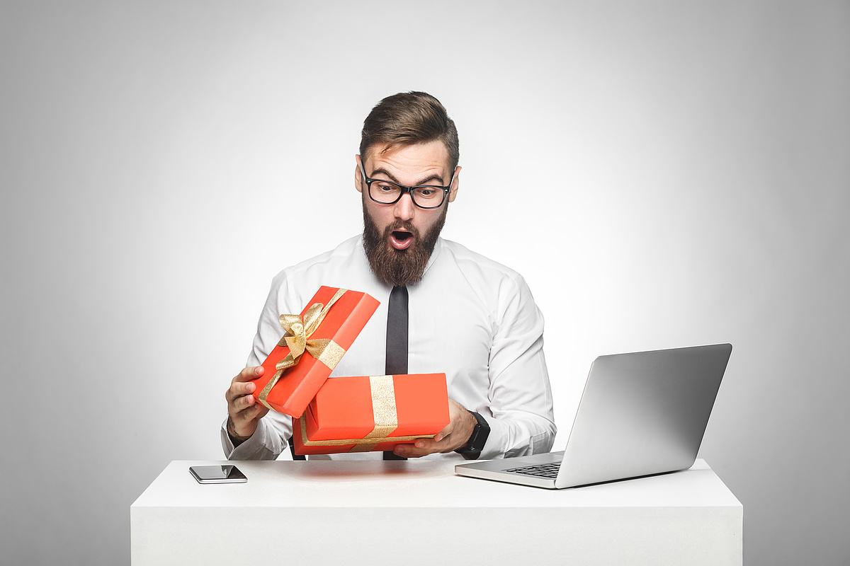 Ein Mann öffnet überrascht ein Geschenk an einem Tisch, neben ihm stehen ein Laptop und ein Smartphone